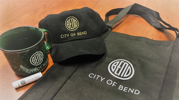 City of Bend swag including a ceramic mug, chapstick, baseball cap and shopping bag.