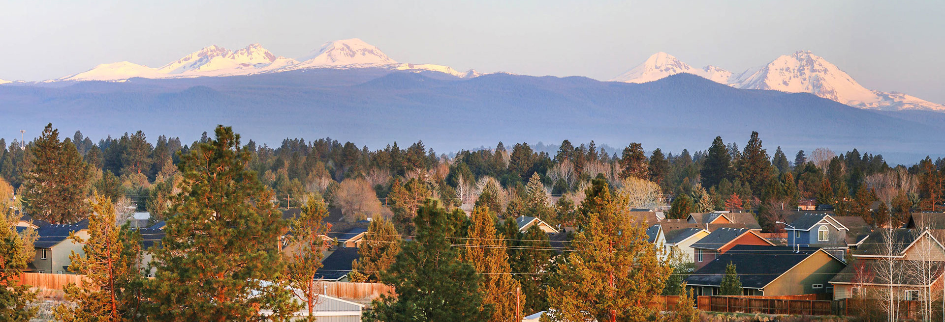 Houses and trees in the foreground, the Three Sisters Mountains and a blue sky in the background