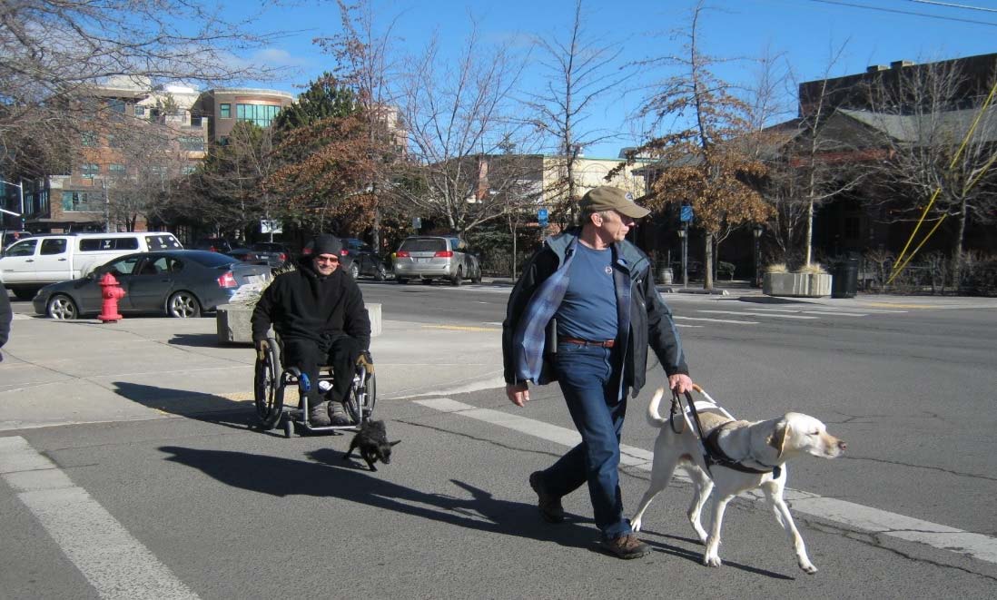 Man in a wheelchair and another man with a service dog cross the street.