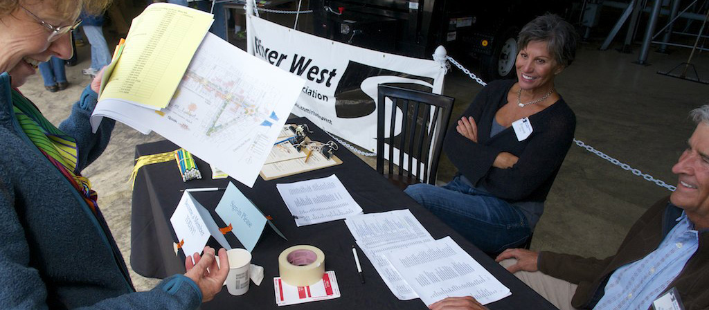 Woman and man sitting behind booth representing a neighborhood association. Another woman looks over some papers in front of them.