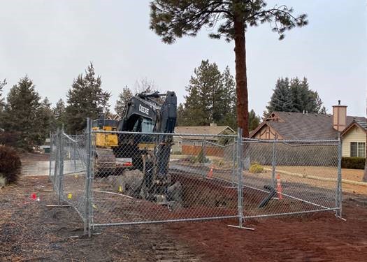 A photo of an excavator excavating within a fenced barricade in a residential neighborhood with pine trees in the background.