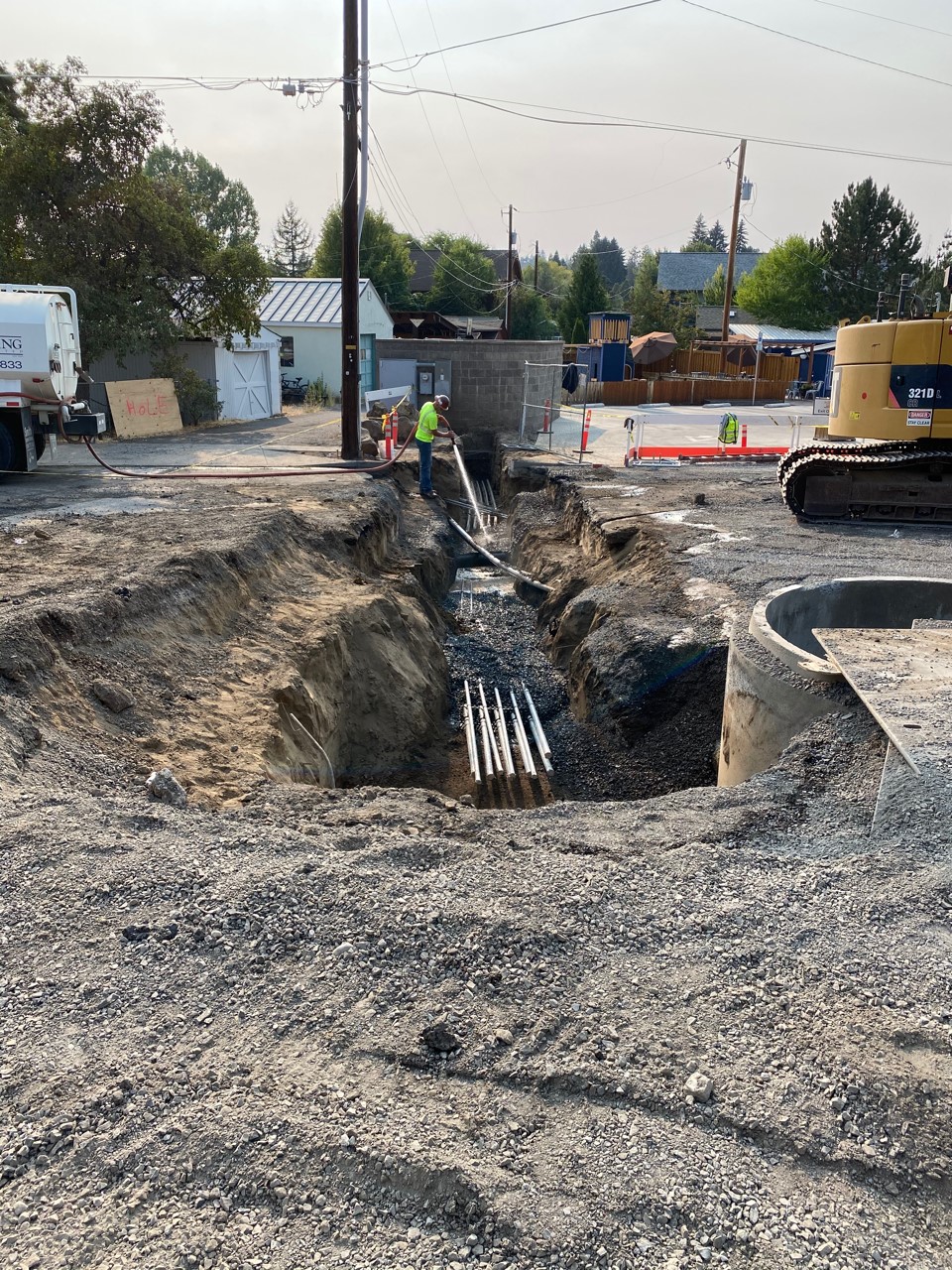 A working crew member spraying water from a water tanker into an excavated trench under NW Riverside St.