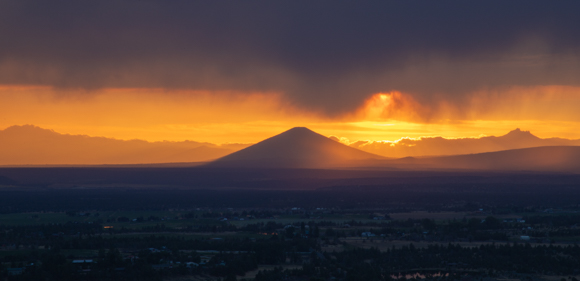 A photo of a mountain against a strip of bright orange sunset between the clouds at dusk.