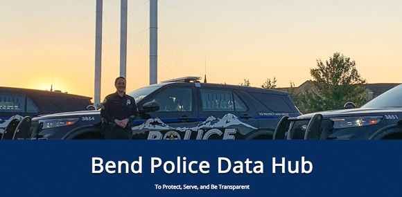 A City of Bend police officer standing in front of a patrol car with the old mill smokestacks in the background