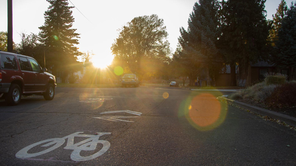 Image of the sun setting over a street with a parked car and a bike lane sign on the asphalt