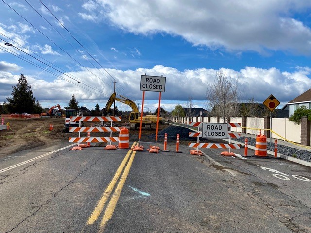 A photo of the construction road closure barricades and cones with work crews and equipment working in the background at the Deschutes Market Road & Butler Market Road roundabout construction site.