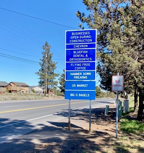 Photo of a blue road sign telling that Chevron, Bluefish Dental & Orthodontics, Flying Frog Coffee, Hammer Down Firearms, US Market, & Big O Bagels are open during construction.