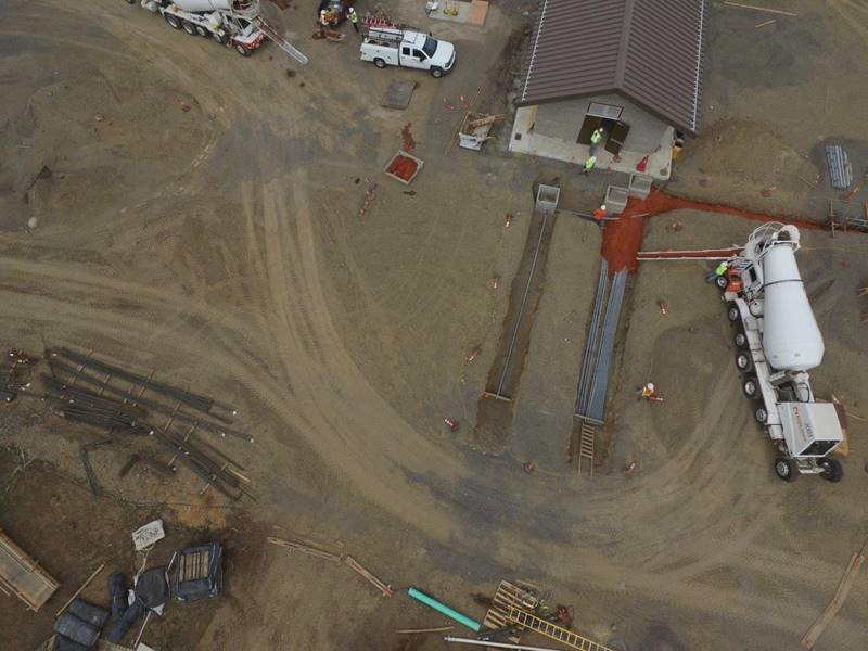 An aerial bird's-eye view of work crews and equipment filling electrical conduit trenches with dyed concrete next to the electrical building.