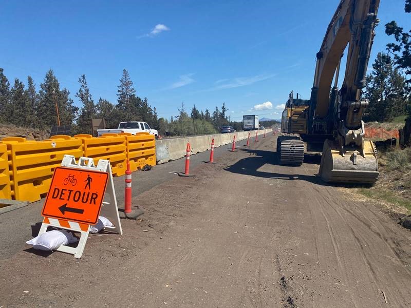 A photo of barricades, cones, and a detour sign along the OR Highway 97 shoulder. There is a large excavator in the shoulder closure, and vehicular traffic is driving by on Highway 97.