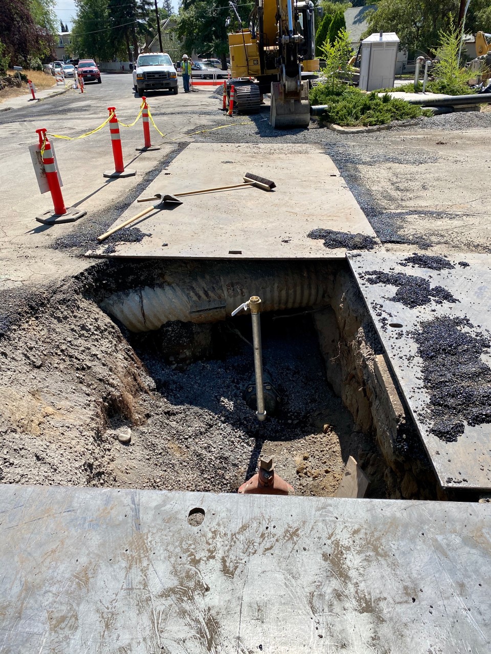 A photo looking over an excavated section of the street showing access to pipes and utility normally hidden below the street. There are work crews, cones, and equiopment in the background.