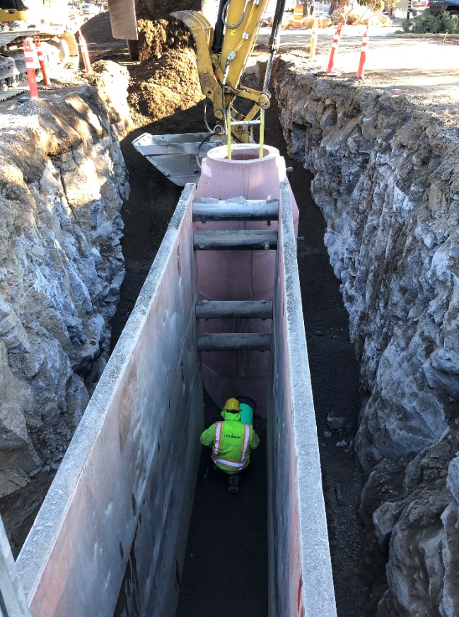 A photo of a working crew member working in a trench box to install a maintenance hole and pipe in a deep trench.