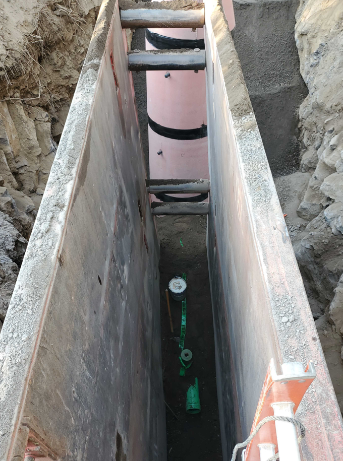 A bird's-eye looking down into a trench box protecting a sewer trench. At the far end of the trench box is a maintenance hole.