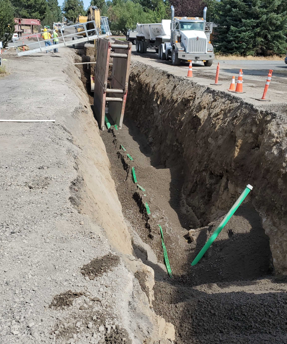 A photo of a sewer trench with a trench wall, buried main pipe with marking tape, and an exposed lateral connection pipe.