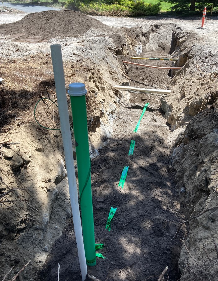 A photo of a sewer lateral trench with other utilities exposed crossing in the open trench. There is a green sewer cleanout & white PVC marker adjacent to it sticking out of the trench. There is also green marker tape laid in the trench.
