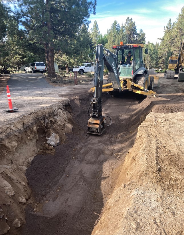 A photo of work crews using a backhoe equipped with a vibratory plate compactor to backfill and compact  the sewer trench.