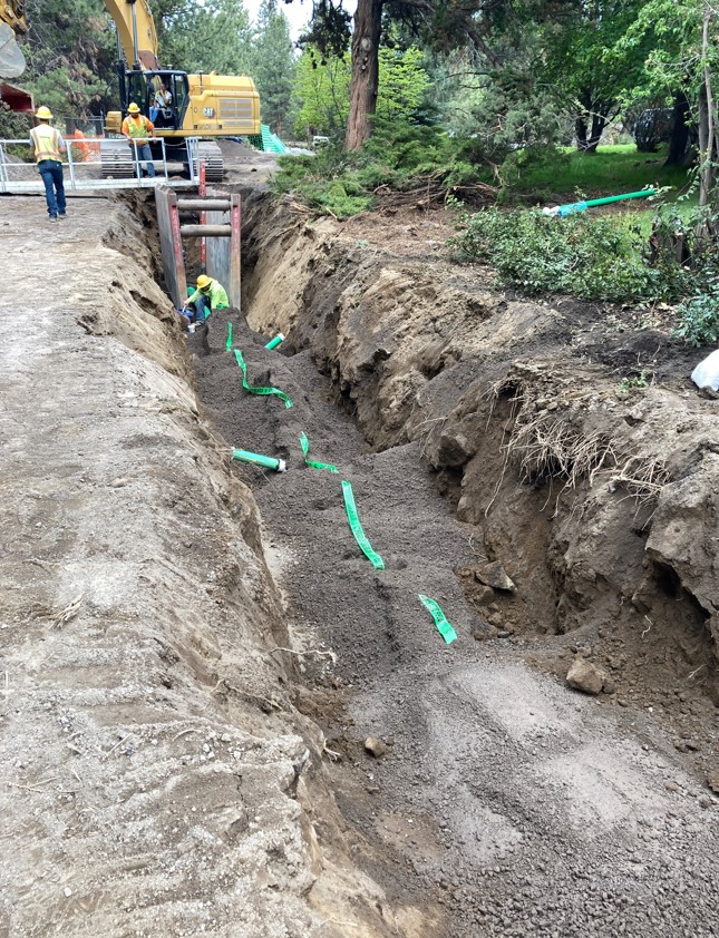 A photo of work crews working at the sewer trench on the southern portion of King Jehu. There is green marking tape laid across the trench. There is a trench box and platform setup at the trench and an excavator working at the edge of the trench.