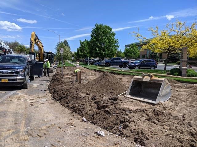 A photo of work crews and excavation equipment preparing for the southeast sidewalks. There is an excavator bucket setting by a mound of dirt in the work area.