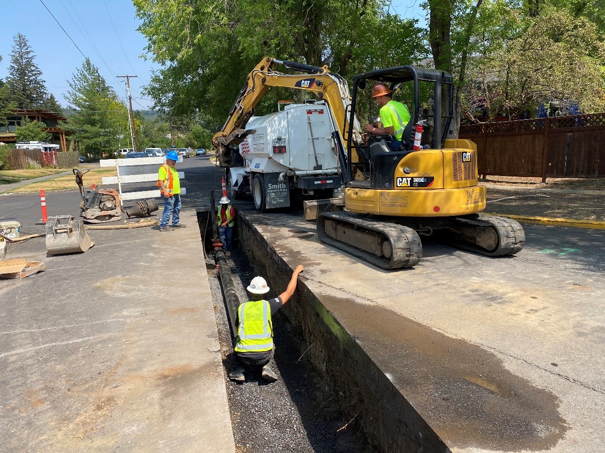 A photo of work crews using a mini-excavator to install the NW Riverside waterline in an excavated trench. There is a tank truck parked in front of the mini-excavator and cones and a barricade marking the construction site.