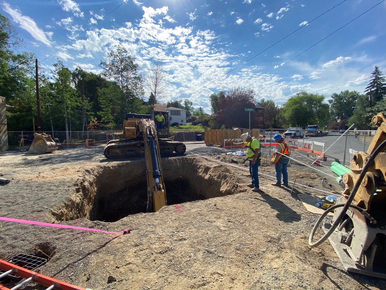 A photo of work crews using excavation equipment and safety harnesses at the we well excavation area. The site is surrounded with cones, fencing and barricades.