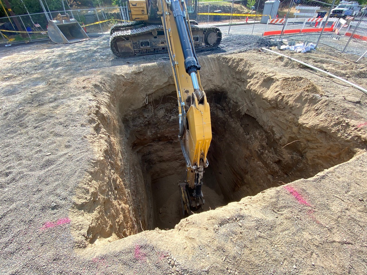A photo looking into the wet well excavation being dug with an excavator's arm extended into the deep trench. There is an excavator bucket in the background and the site is surrounded with barracades, caution tape, cones, and fencing.