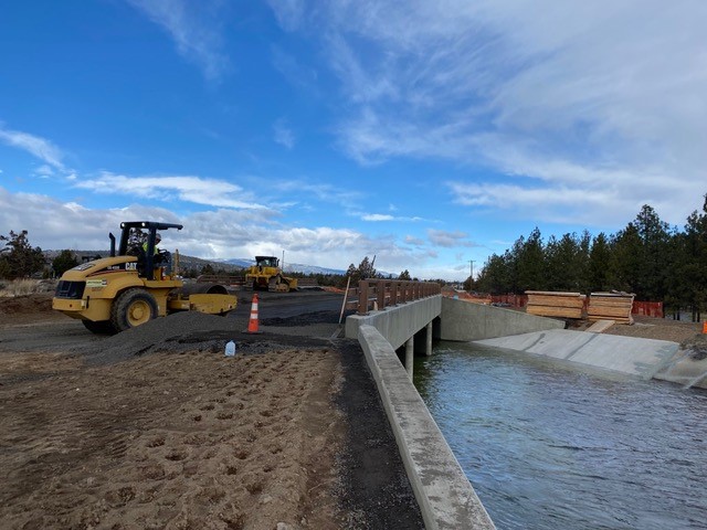 A photo of work crews working with a compactor and dozer to continue construction of the Empire crossing, as irrigation water flows in the canal below the crossing structure.
