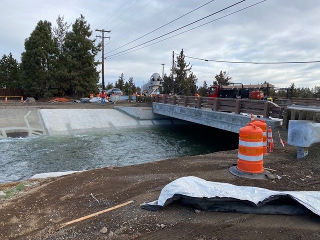 A photo of work crews and equipment pouring sidewalks on the NE Purcell bridge, as irrigation water flows in the canal under the bridge.
