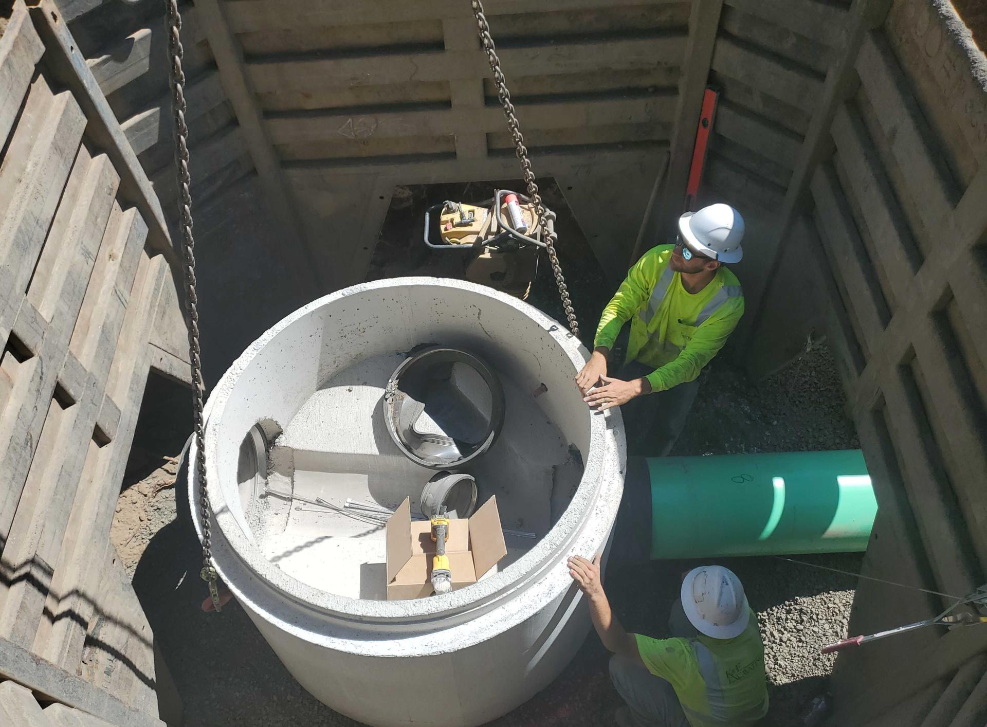A photo of two construction crew members working to carefully lower a sewer maintenance hole base section into a trench.