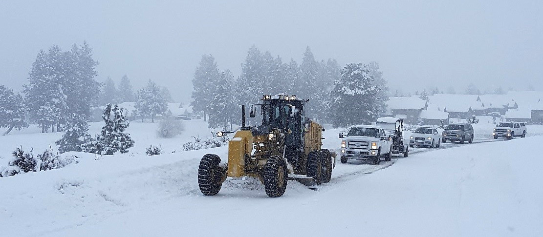 Grader plowing snow off of the road during snowstorm with line of cars behind.