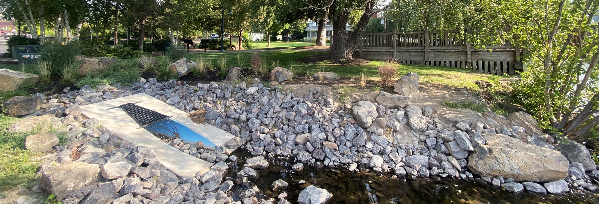 Storm drainpipe jetting into a riverbed. Rocks and trees surround the pipe.