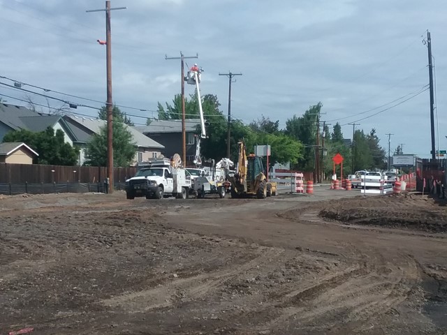 A photo of work crews performing utility work on NE Purcell Blvd. and NE Butler Market Rd. Crew members are using a cherry picker/aerial lift to work on a utility pole. There are barricades, signs, and cones at the road closure on NE Purcell Blvd.