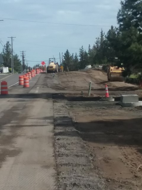 A photo of work crews using equipment for road base, curb, and sidewalk preparations on NE Purcell Blvd. There are cones and road signs along the shoulder of NE Purcell Blvd.