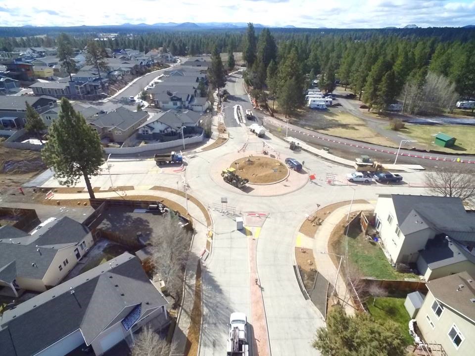 An aerial photo of the Murphy/Brosterhous roundabout looking south over work crews, equipment, surrounding neighborhoods, and a landscape of pine trees and hills in the background.