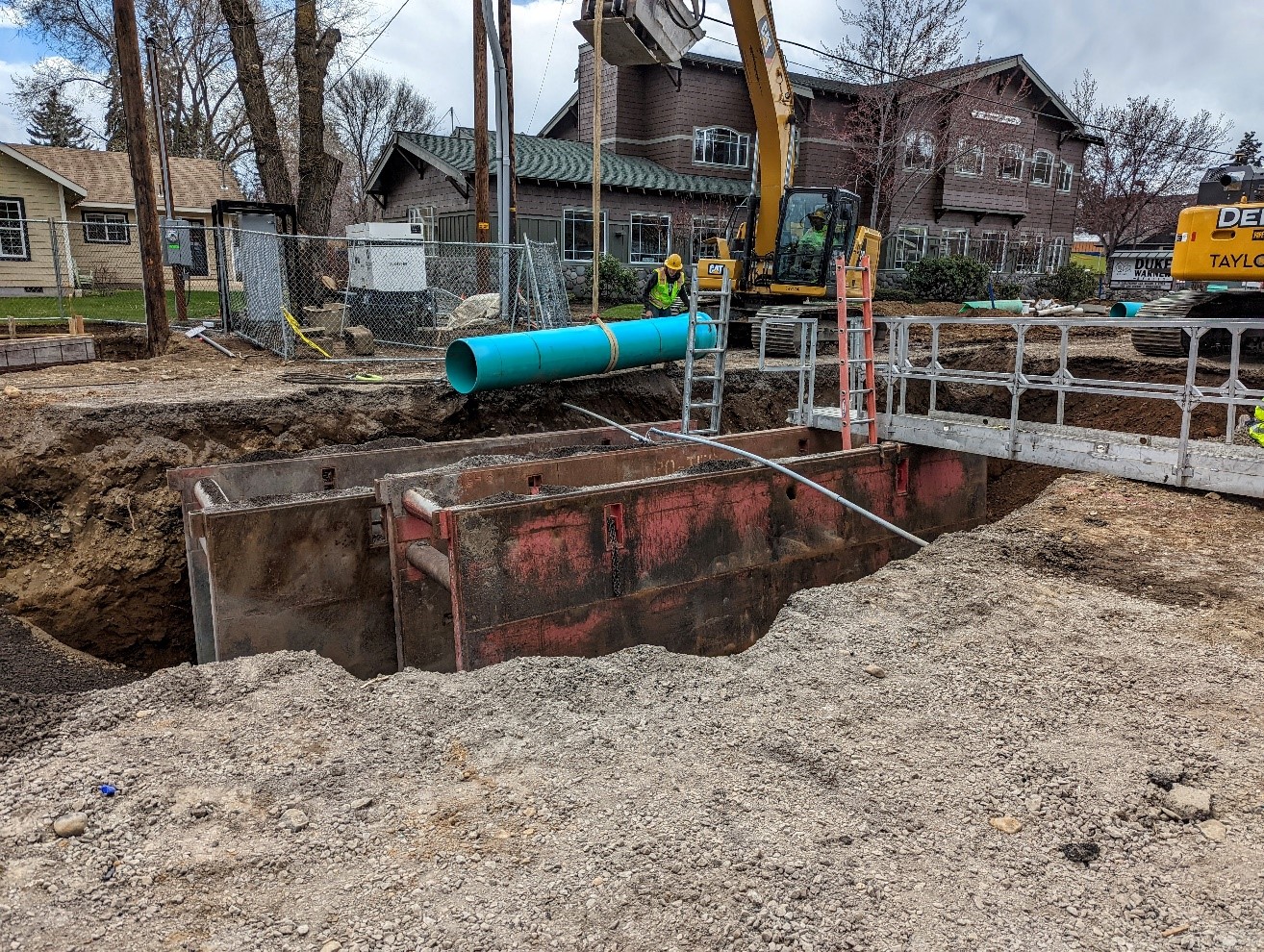 A photo of work crews using equipment to install new pipe in a deep trench. There is a trench shield, ladders, and a bridge with rails for safe access to the trench.