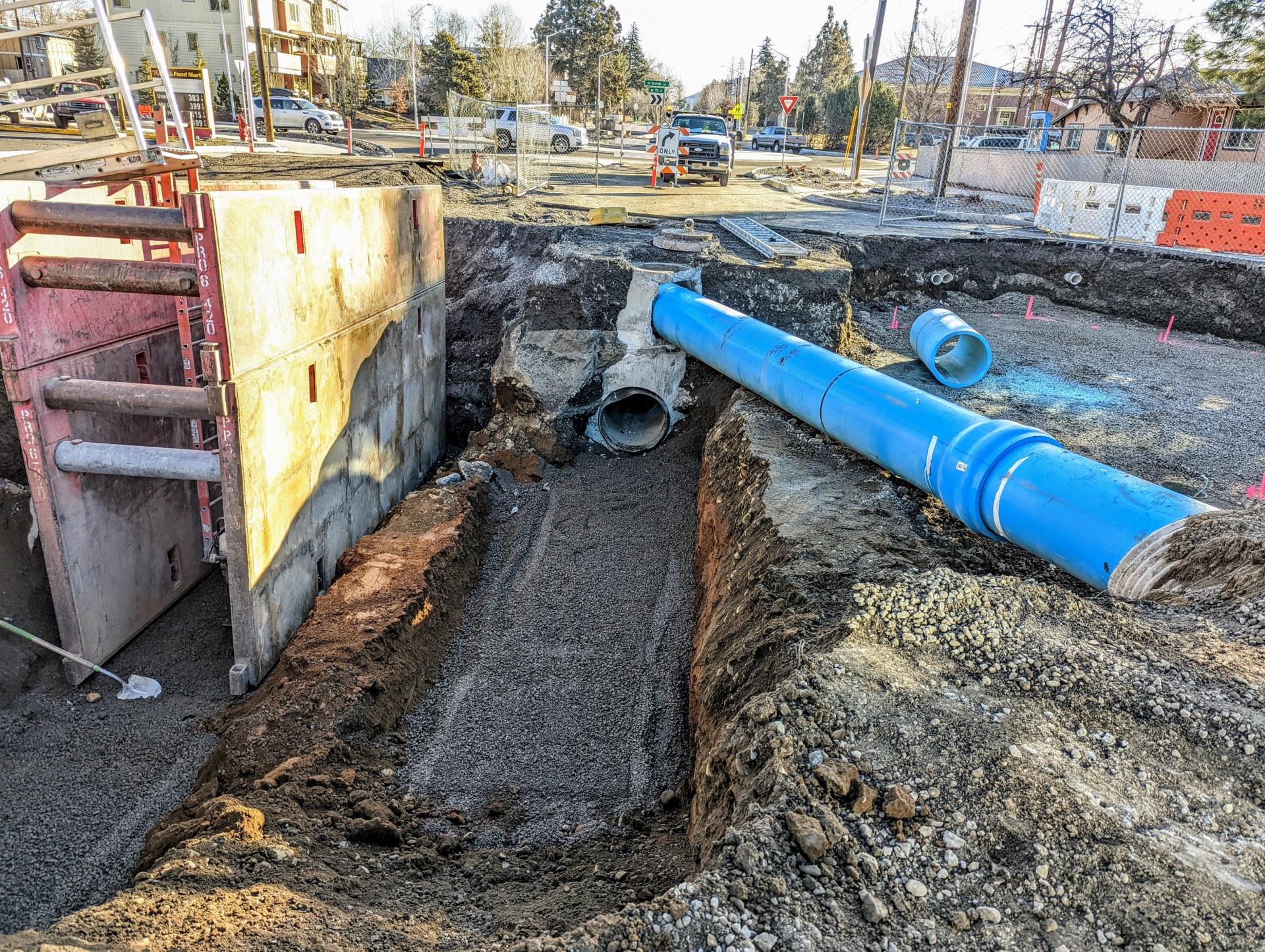 A photo of an excavated trench with exposed utility lines and a shoring device setup in the trench. There is fencing, cones, and barricades setup around the excavated site for safety.