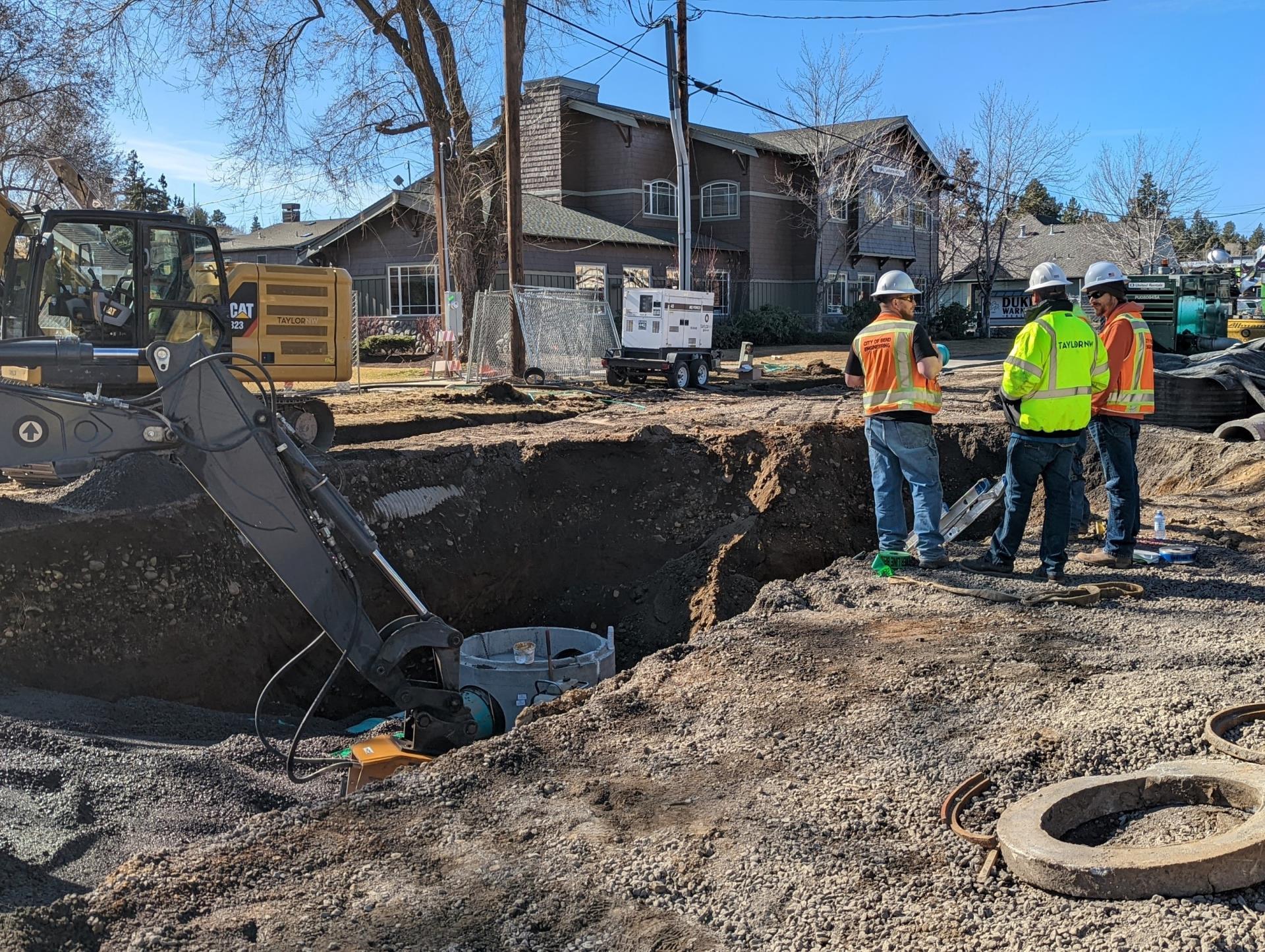 A photo of two City of Bend Inspectors wearing orange safety vests, white hardhats, and sunglasses conversing with a work crew member wearing a neon green reflective jacket and white hardhat standing next to an excavated trench where stormwater pipes are exposed.