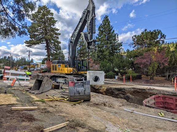 A photo of work crews using a excavator rigged with a sling to lower a new stormwater structure into a trench on Newport Ave.