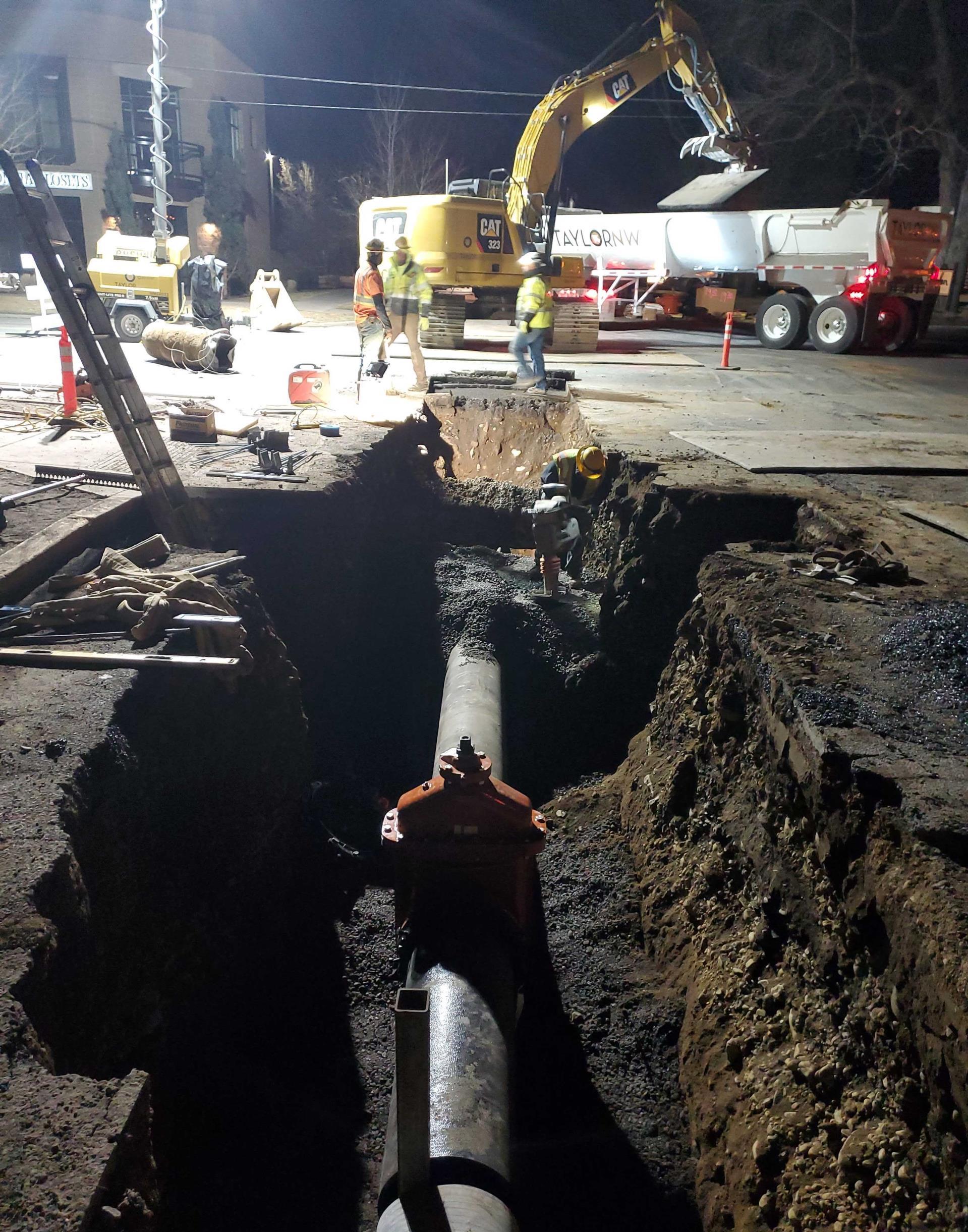 A photo of work crews performing night waterline work at Newport Ave. & 10th St. The new waterline is installed in the open trench and there are cones, lighting, and large equipment on the project site.