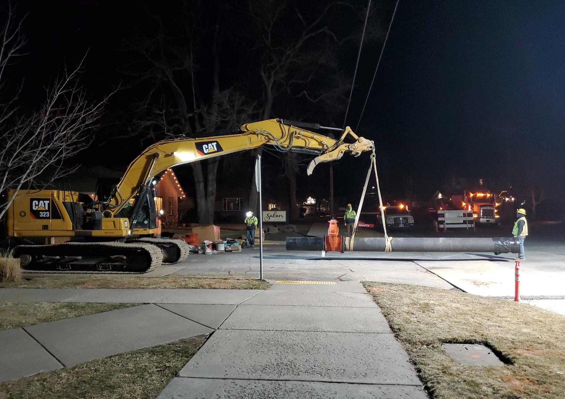 A photo of a night working crew using a hydraulic excavator to move and install a new waterline at Newport Ave. & 10th St.