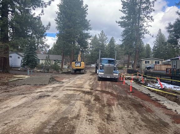 A photo of the removal of existing roadway along McMullin Drive. There are cones and caution tape surrounding an excavated trench along the right side of the roadway. There is a truck and excavator on the roadway.