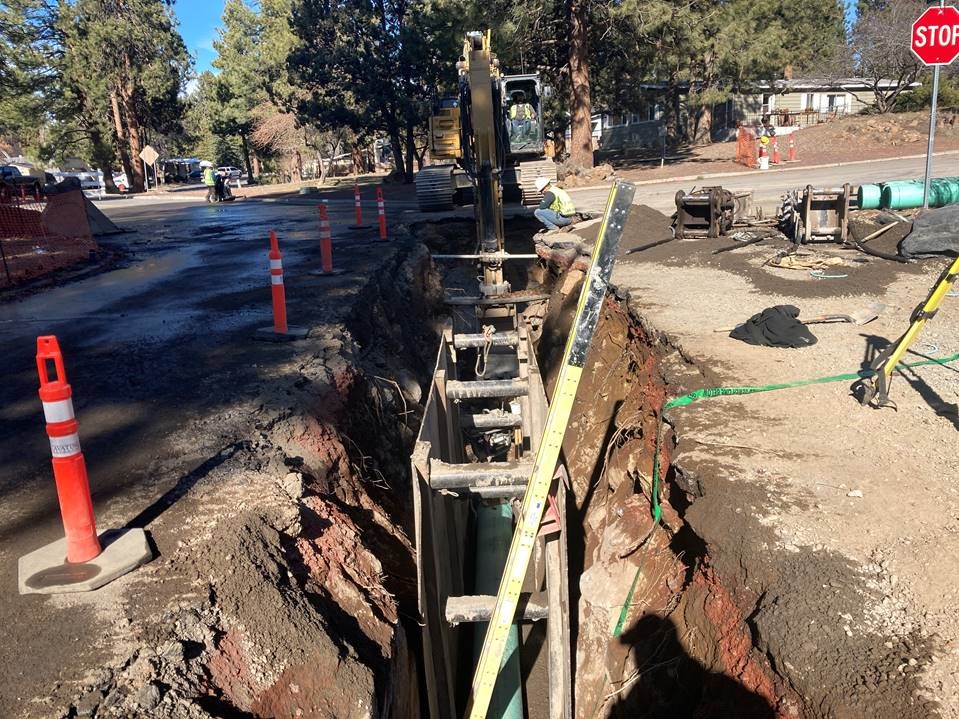 A photo of the sewer main installation in an excavated trench in McMullin Drive, near Mahogany Street. There are orange cones along the left-side edge of the trench and a ladder poking out of the right-side edge of the trench.