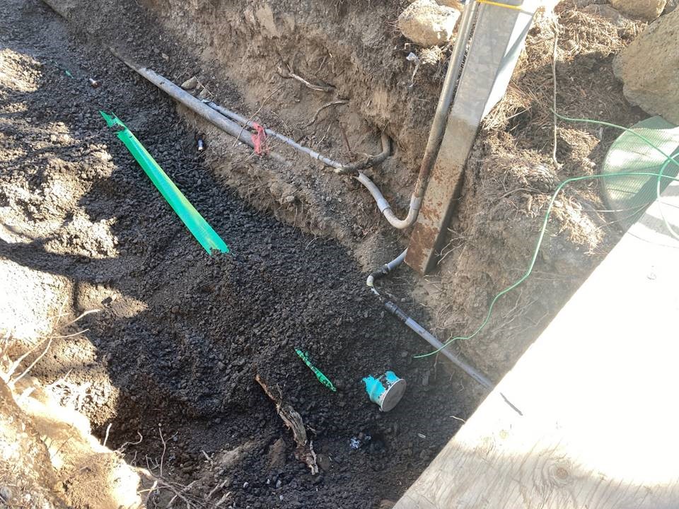 A photo of an excavated trench and exposed utility lines. There is a strip of green marker tape partially exposed in the trench.