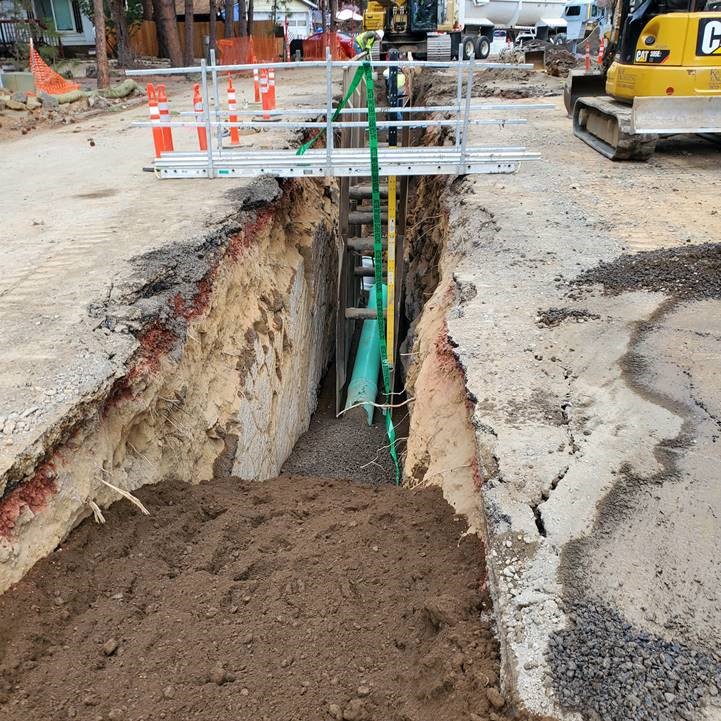 A photo of the sewer main installation in an open trench in McMullin Drive. There is a trench bridge with railing in place over the trench, and cones along the edge of the trench.