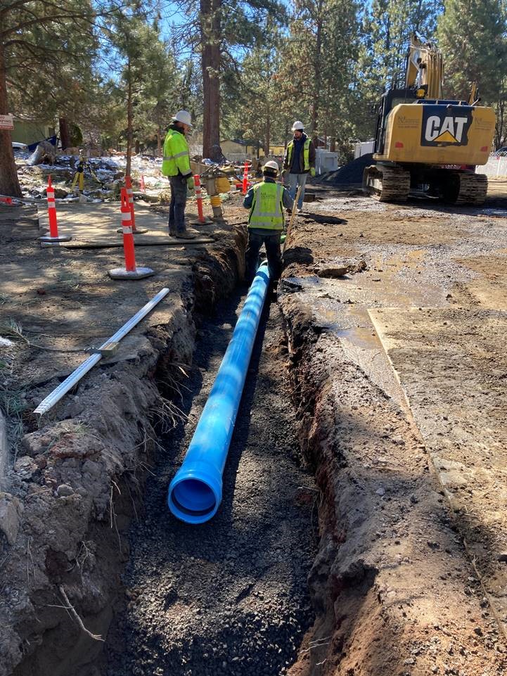 Work crews are installing new blue storm pipe in an excavated trench along McMullin Drive. There are orange cones along the left side of the trench, and a yellow excavator on the right side of the trench.