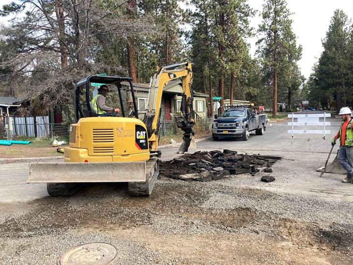 a photo of work crews using a mini excavator to break up the roadway and tie in the final service line to the new main along Mahogany Street.