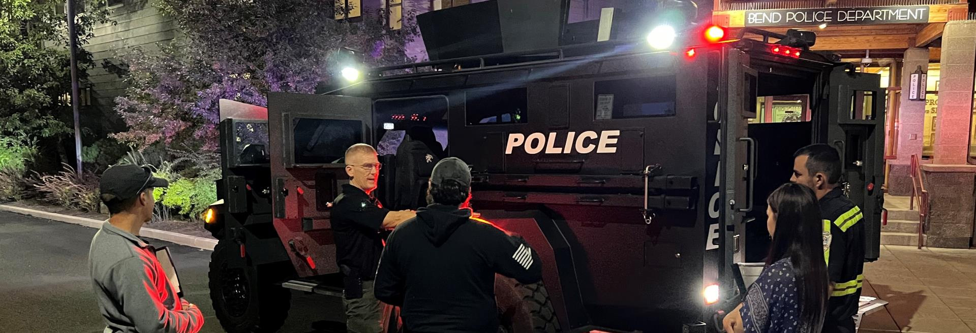 Police officers and community members outside a building next to an armored police vehicle.