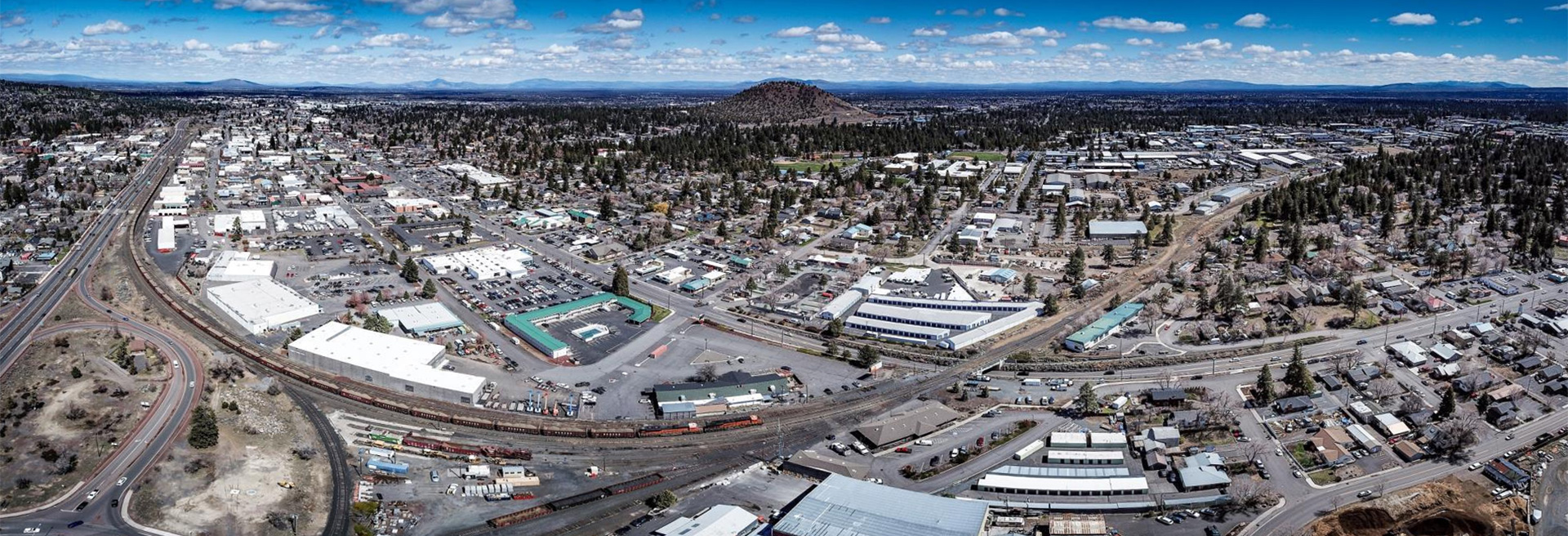 Overhead shot of Bend's Core Area looking northeast. Roads, commerical buildings, sidewalks, cars. Pilot Butte is in the mid-ground, partly cloudy skies.