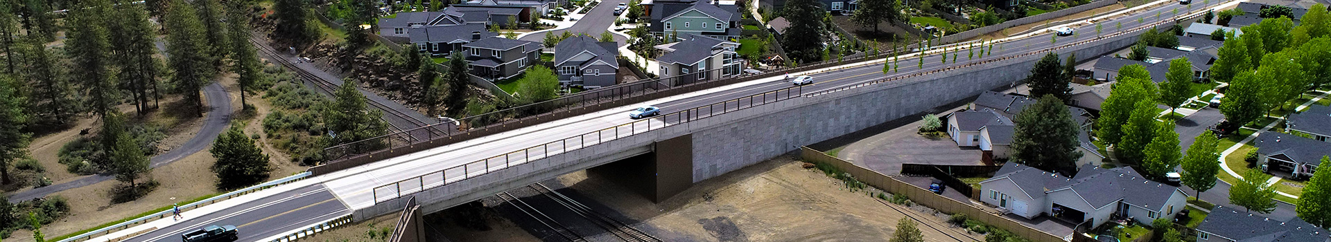 Bridge under construction going over the top of railroad tracks. Houses and trees surround the bridge.