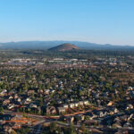 City of Bend looking northeast from a drone. City buildings, houses, streets, and trees. Pilot Butte in the mid-ground. Blue skies and hills in the background.