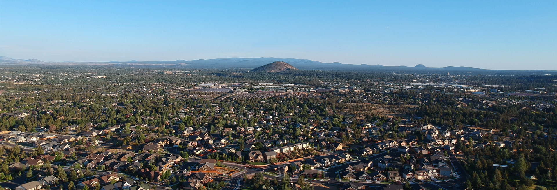 City of Bend looking northeast from a drone. City buildings, houses, streets, and trees. Pilot Butte in the mid-ground. Blue skies and hills in the background.