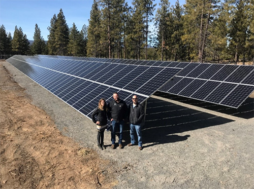 Large solar panels on the ground with two men and one woman standing in front of them. Trees and blue sky in the background.